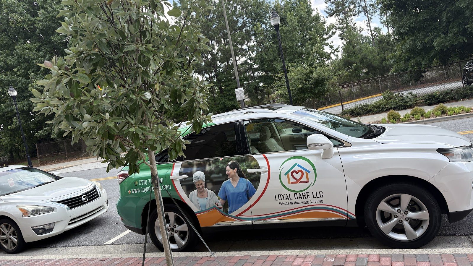 A white SUV wrapped in the Loyal Care LLC logo with green and orange panels, parked on a tree-lined street